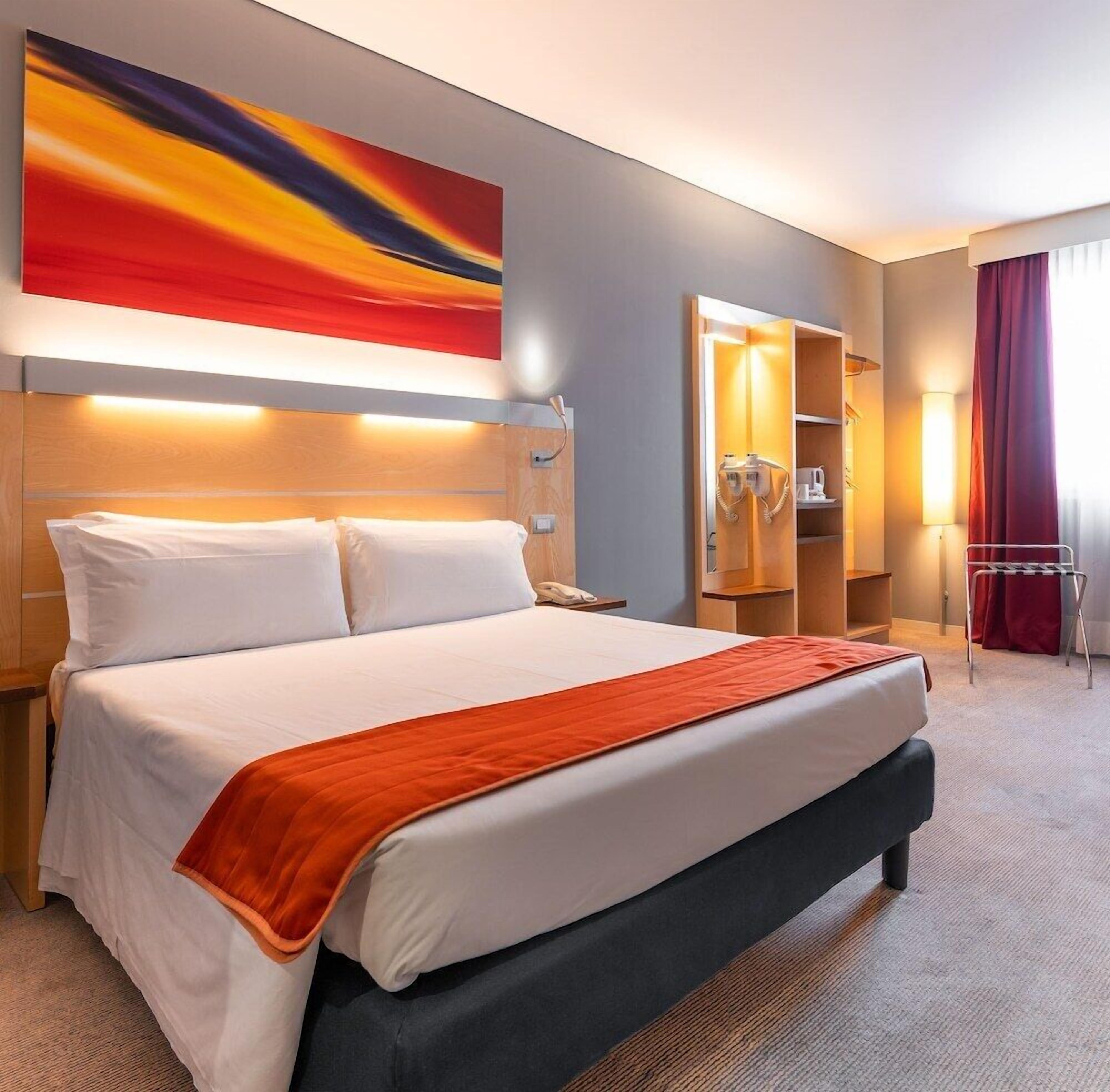 A hotel room with a large bed, a headboard with integrated lighting, a bedside table with a phone, a wardrobe with a mirror, and a red curtain.