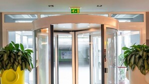 A revolving door with glass panels, two large potted plants, and a green exit sign above. - Bes Hotel Bergamo West (Mozzo)