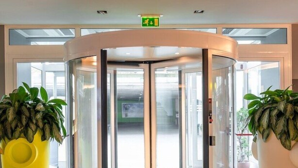 Une porte tournante avec des panneaux de verre, deux grands pots de plantes et un panneau de signalisation vert indiquant la sortie, placé au-dessus.