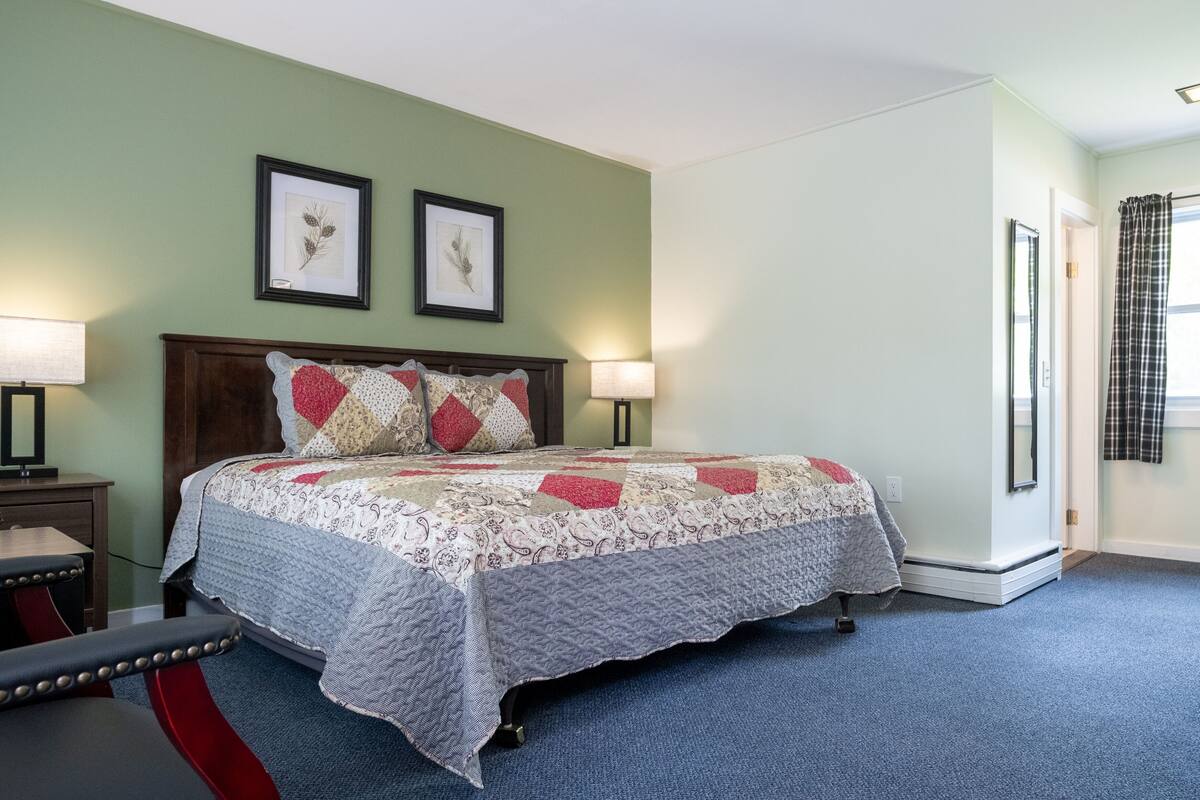 A bedroom with a bed, two framed botanical prints on the wall, a nightstand with a lamp, and a window with curtains.
