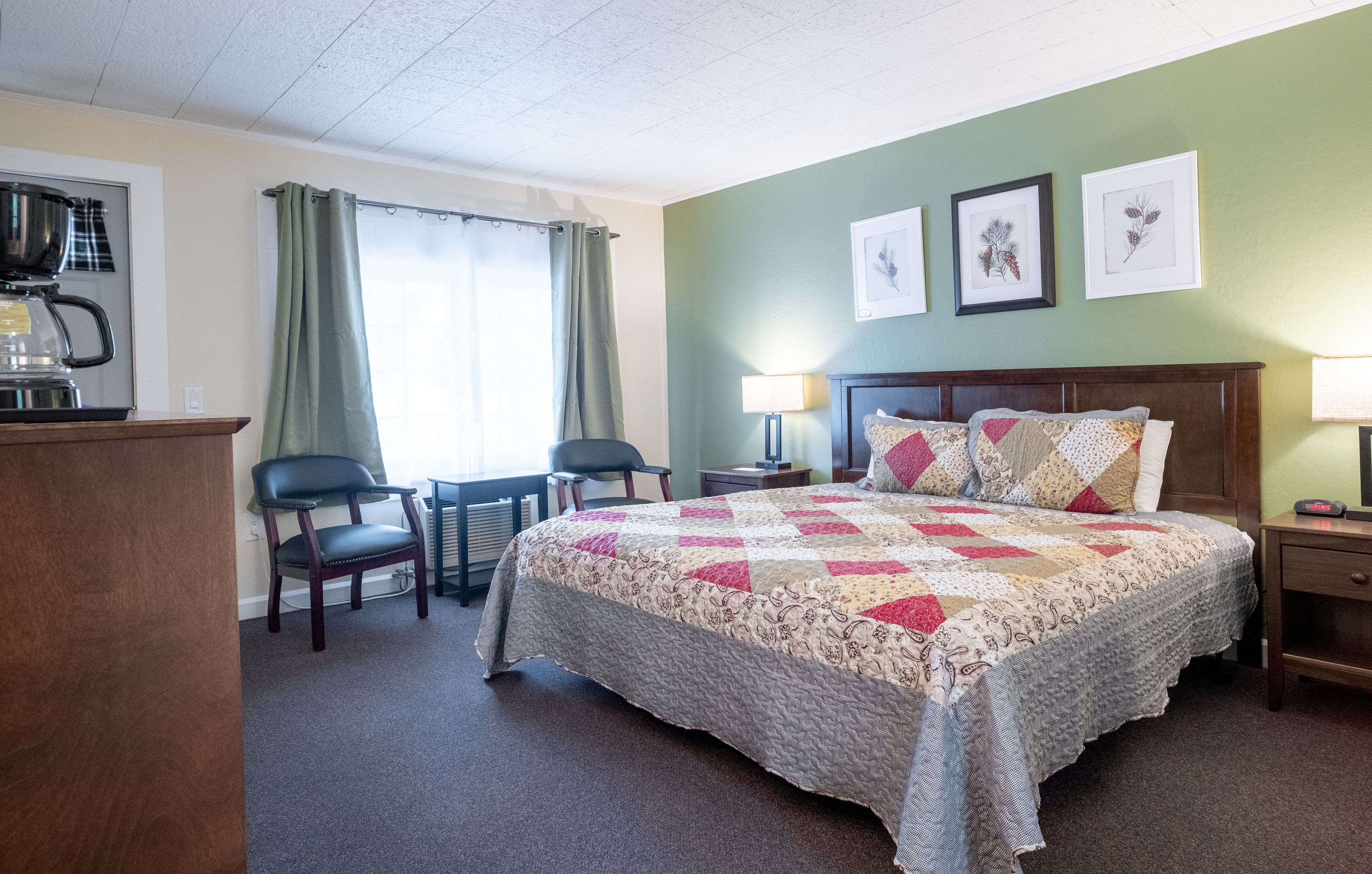 A hotel room with a bed, a desk with a coffee maker, two chairs, a lamp, and framed botanical prints on the wall.