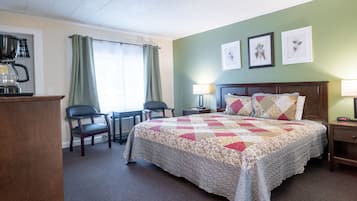 A hotel room with a bed, a desk with a coffee maker, two chairs, a lamp, and framed botanical prints on the wall.