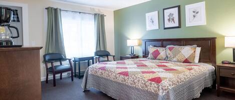 A hotel room with a bed, a desk with a coffee maker, two chairs, a lamp, and framed botanical prints on the wall.