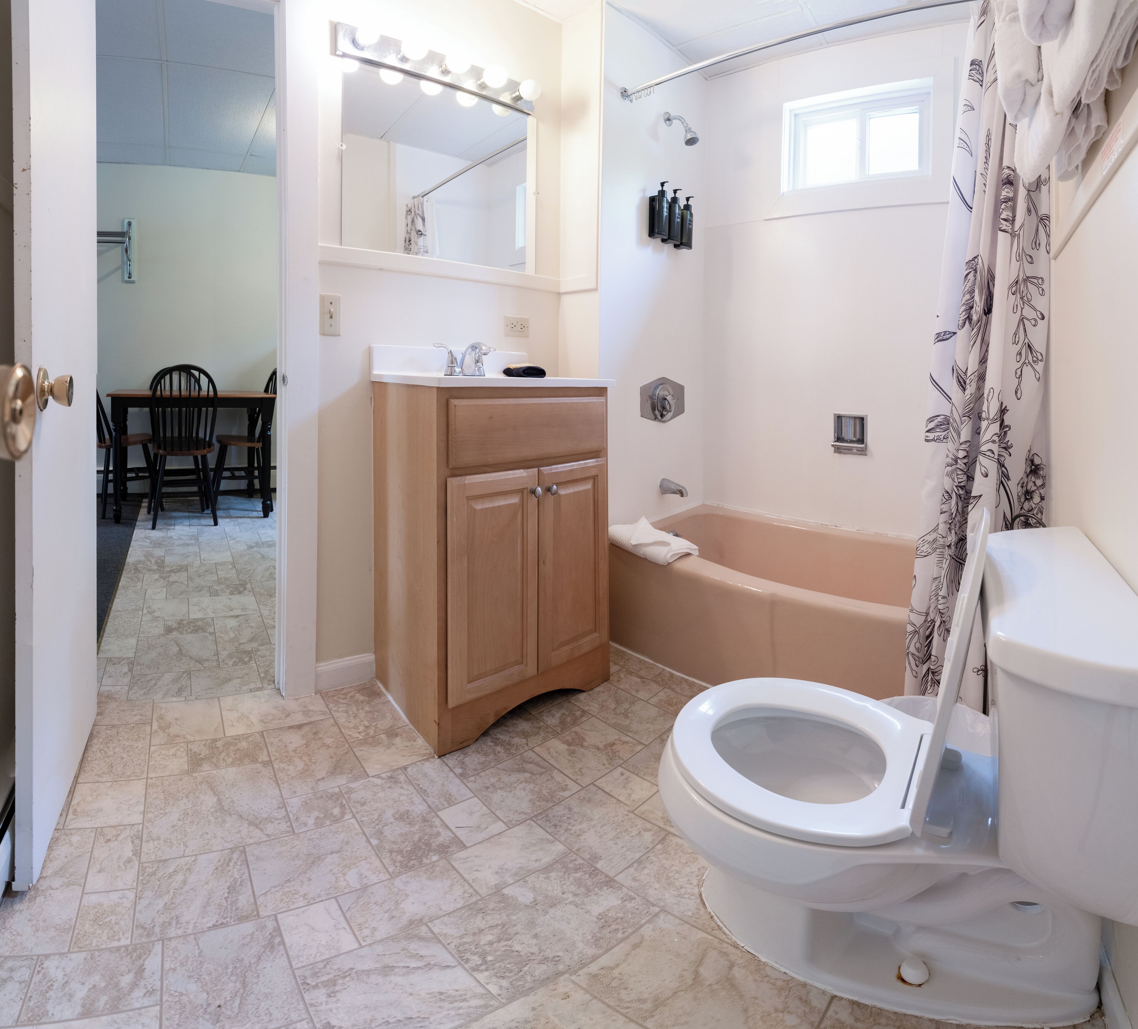 A bathroom with a toilet, bathtub, and sink. There is a vanity with a mirror and lights above the mirror. The floor is tiled, and there is a shower curtain with a floral pattern.