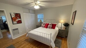 Une chambre avec un lit en métal blanc, un ventilateur de plafond, une fenêtre avec des stores, une table de chevet avec une lampe et un tableau encadré au mur.