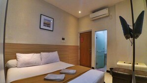 Habitación de hotel con cama, cabecera de madera, un cuadro enmarcado en la pared, unidad de aire acondicionado y mesita de noche con lámpara.