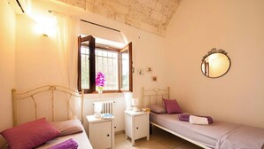 Quarto com duas camas, um radiador, uma mesa de cabeceira, uma janela com cortinas, um espelho e um pequeno vaso com flores.