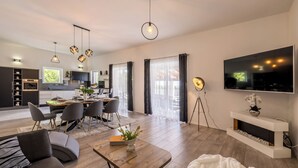 Uma sala de estar moderna com um sofá, mesa de centro e televisão. Há uma área de jantar com cadeiras e mesa. A cozinha é visível ao fundo.