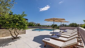 Uma área de piscina no terraço com espreguiçadeiras e um guarda-sol, cercada por um muro de pedra e vegetação.