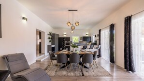 Uma sala de jantar moderna com uma mesa de madeira posta para quatro pessoas, rodeada por cadeiras cinzentas. Uma cozinha planejada fica ao fundo, juntamente com um sofá cinza e uma grande janela com cortinas.
