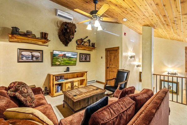 A cozy living room with a wooden ceiling, a mounted buffalo head, a wooden coffee table, a leather sofa, and a television.