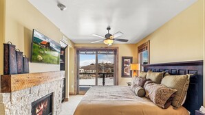 A bedroom with a stone fireplace, a bed with patterned bedding, a ceiling fan, and a large window overlooking a scenic view. - Edgemont 2507 Luxury On Mountain Condo With Ski-In/Ski-Out Access! (Steamboat Springs)