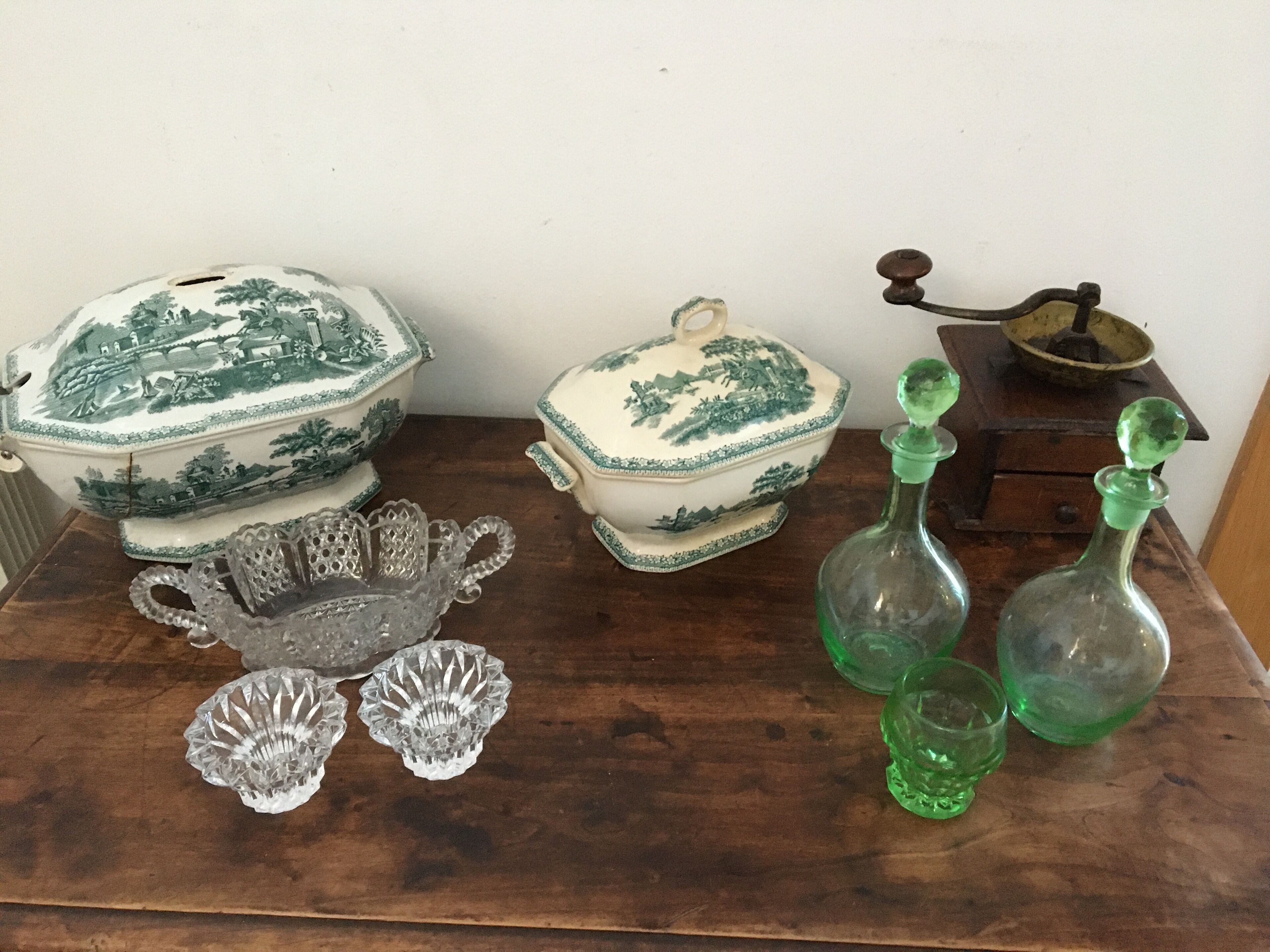 Une table en bois, ornée d’une vaisselle en porcelaine ancienne, de verres en cristal et de bouteilles en verre vert.
