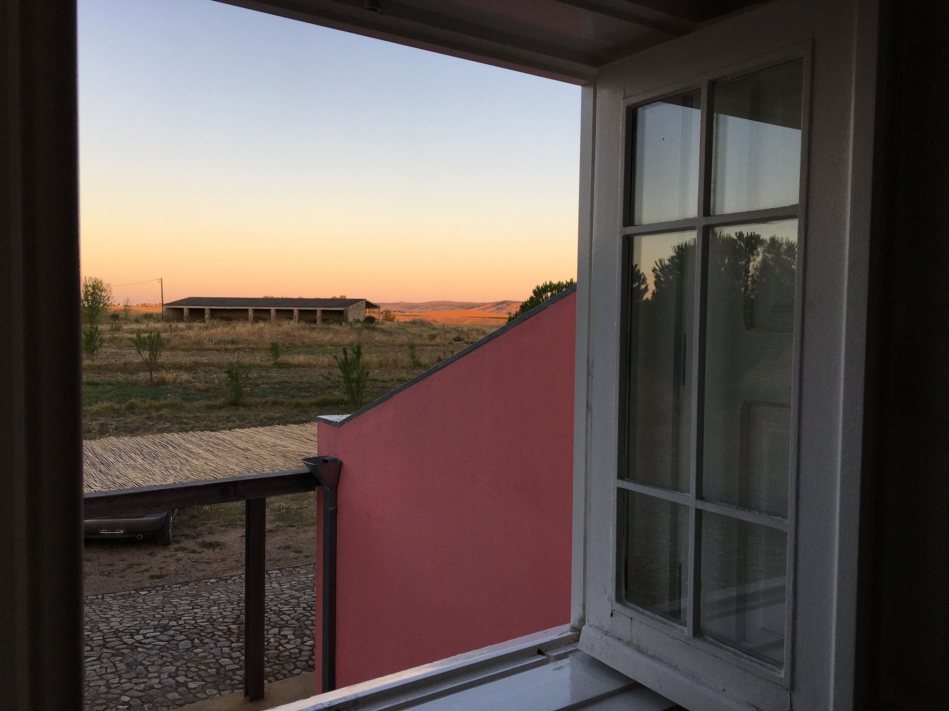 Une vue depuis une fenêtre donnant sur un bâtiment à la façade rouge, une zone pavée avec un banc et un autre bâtiment situé au loin.