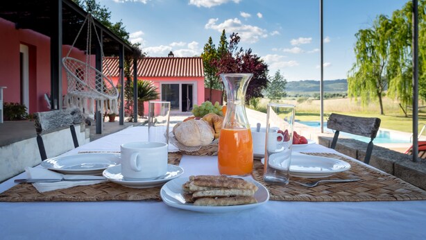 A table set for breakfast with plates, cups, and a bottle of orange juice, overlooking a pool and a house.