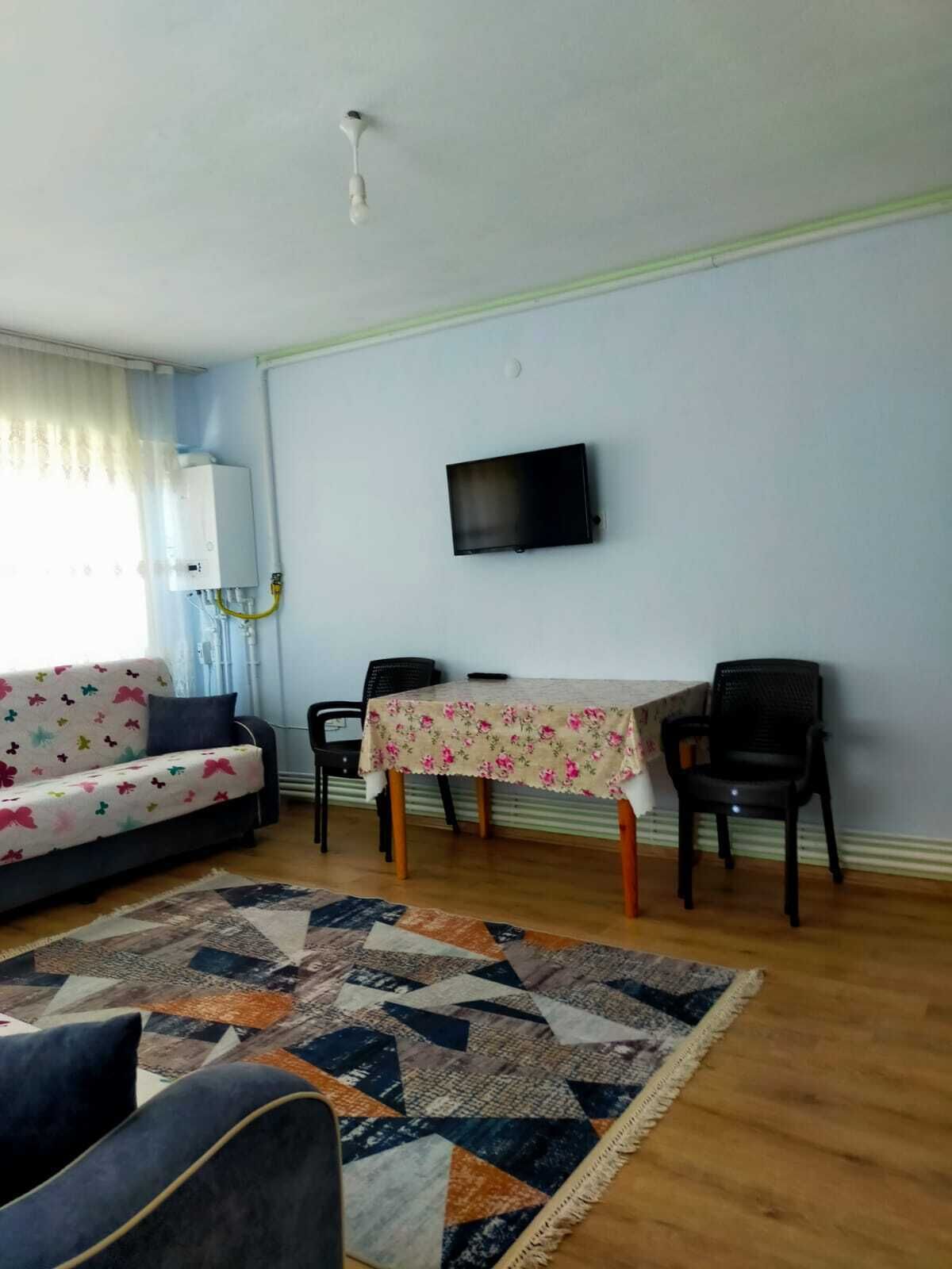 A living room with a sofa, a dining table with two chairs, a TV mounted on the wall, and a patterned rug.