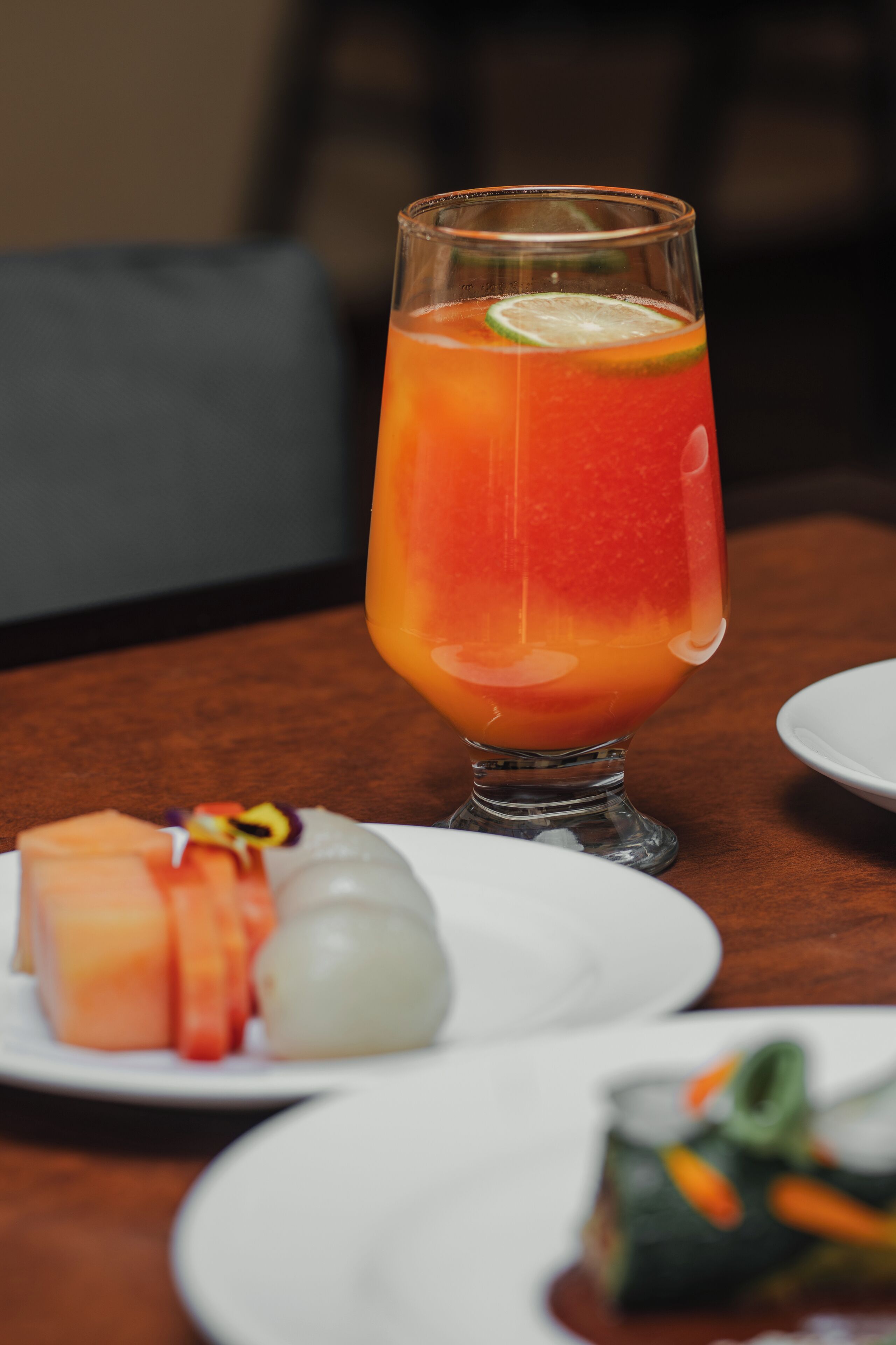 A glass of orange-colored drink with a lime slice on a table with plates of food.