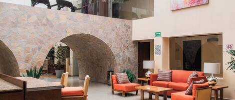 Un vestíbulo amplio con techo alto, arcos de piedra, una zona de estar central con sofás naranjas y un gran cuadro abstracto en la pared.