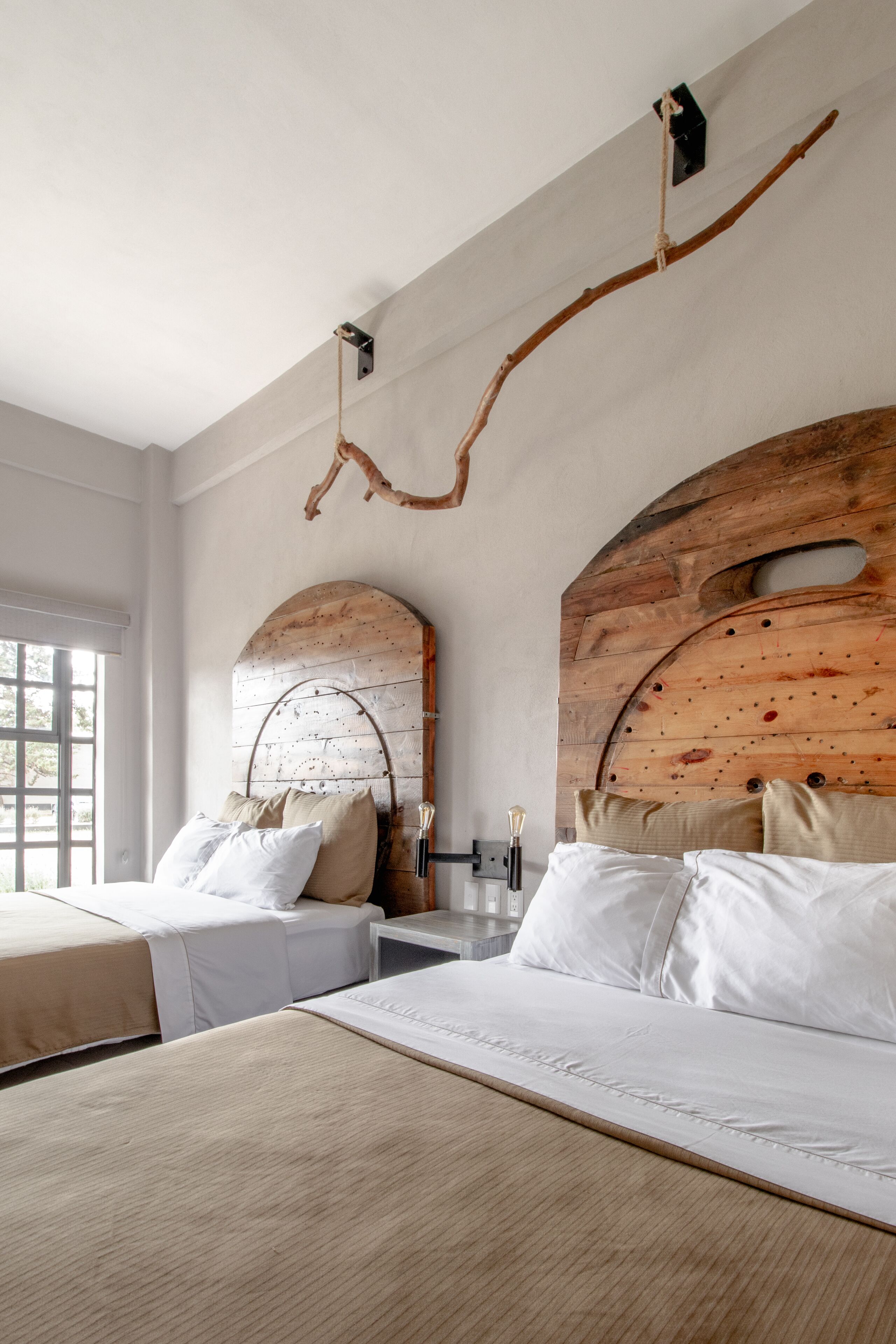 Habitación de hotel con dos camas, cabeceros de madera, un ventanal grande y un adorno colgante de ramas.