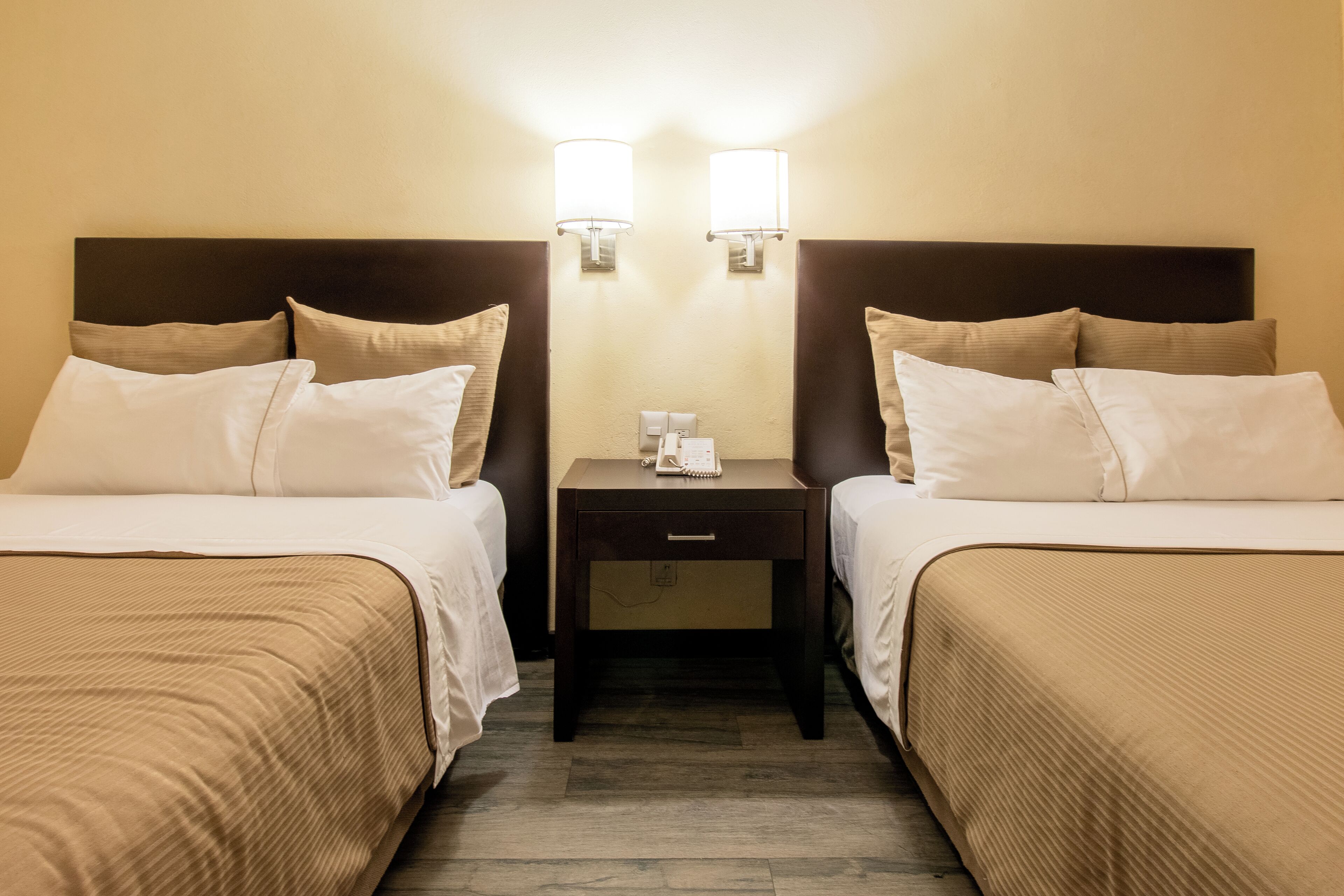 A hotel room with two beds, each with a headboard, a bedside table with a lamp, and a phone.