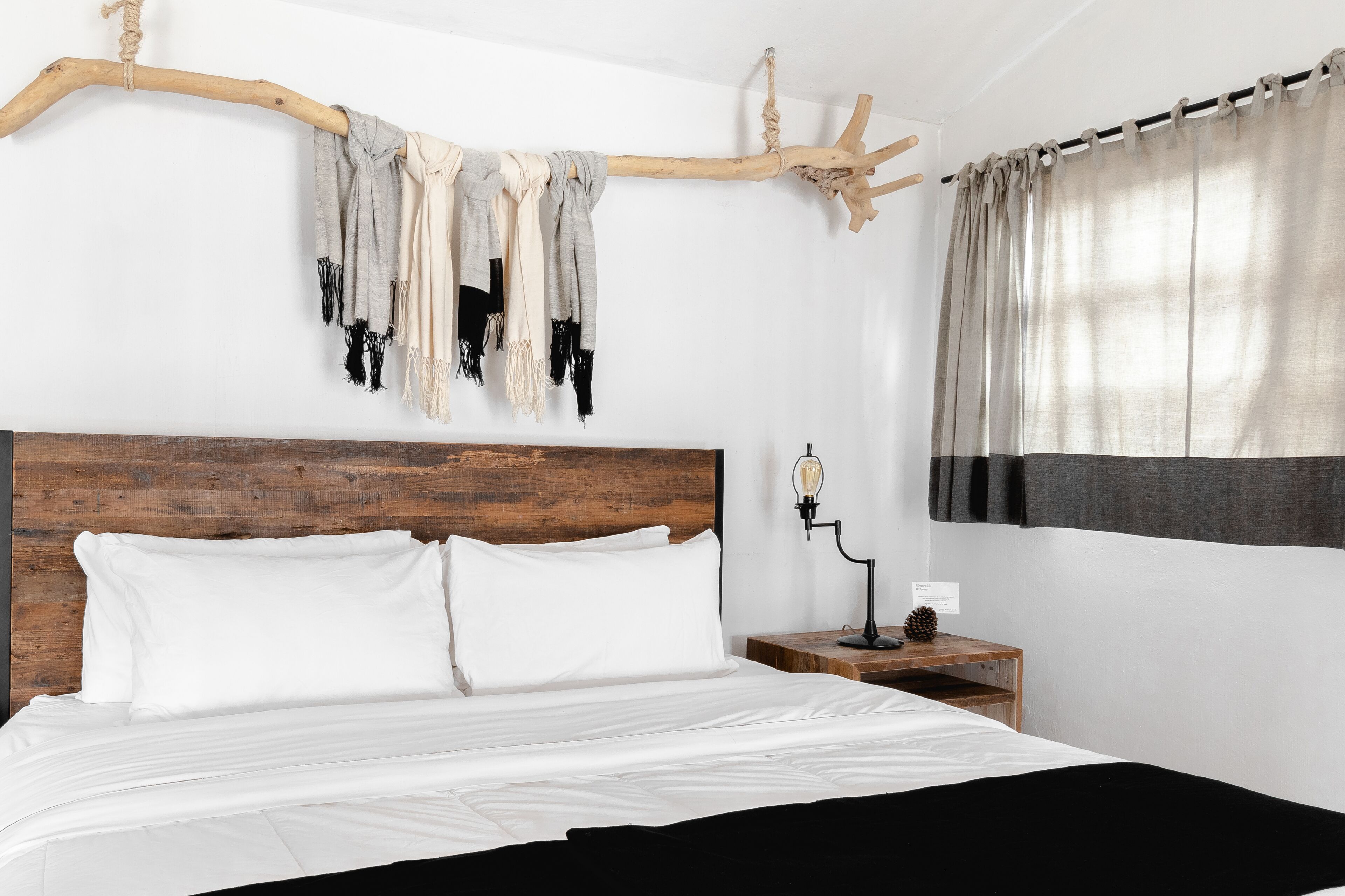 Une chambre à coucher avec une tête de lit en bois, un lit recouvert de draps blancs, une table de chevet avec une lampe et un rideau à la fenêtre.