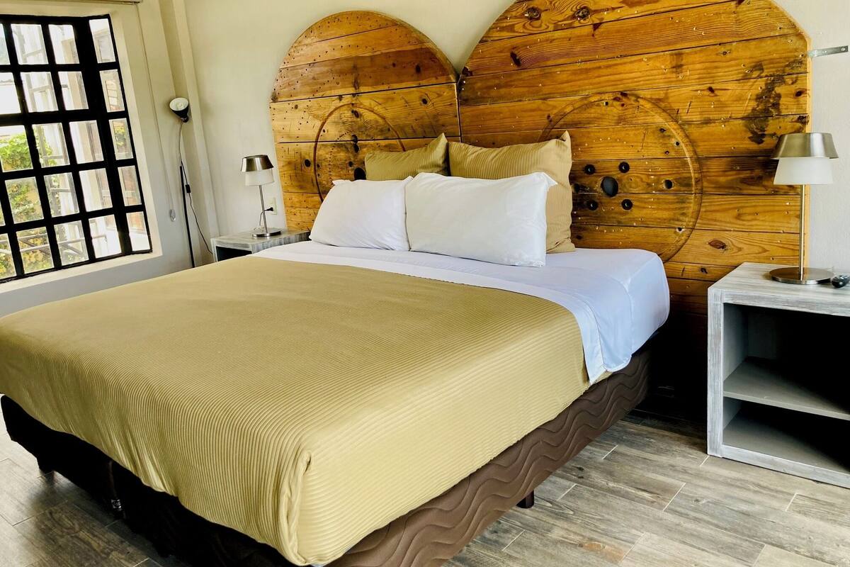 A bed with a wooden headboard, a bedside table with a lamp, and a window with a view of greenery.