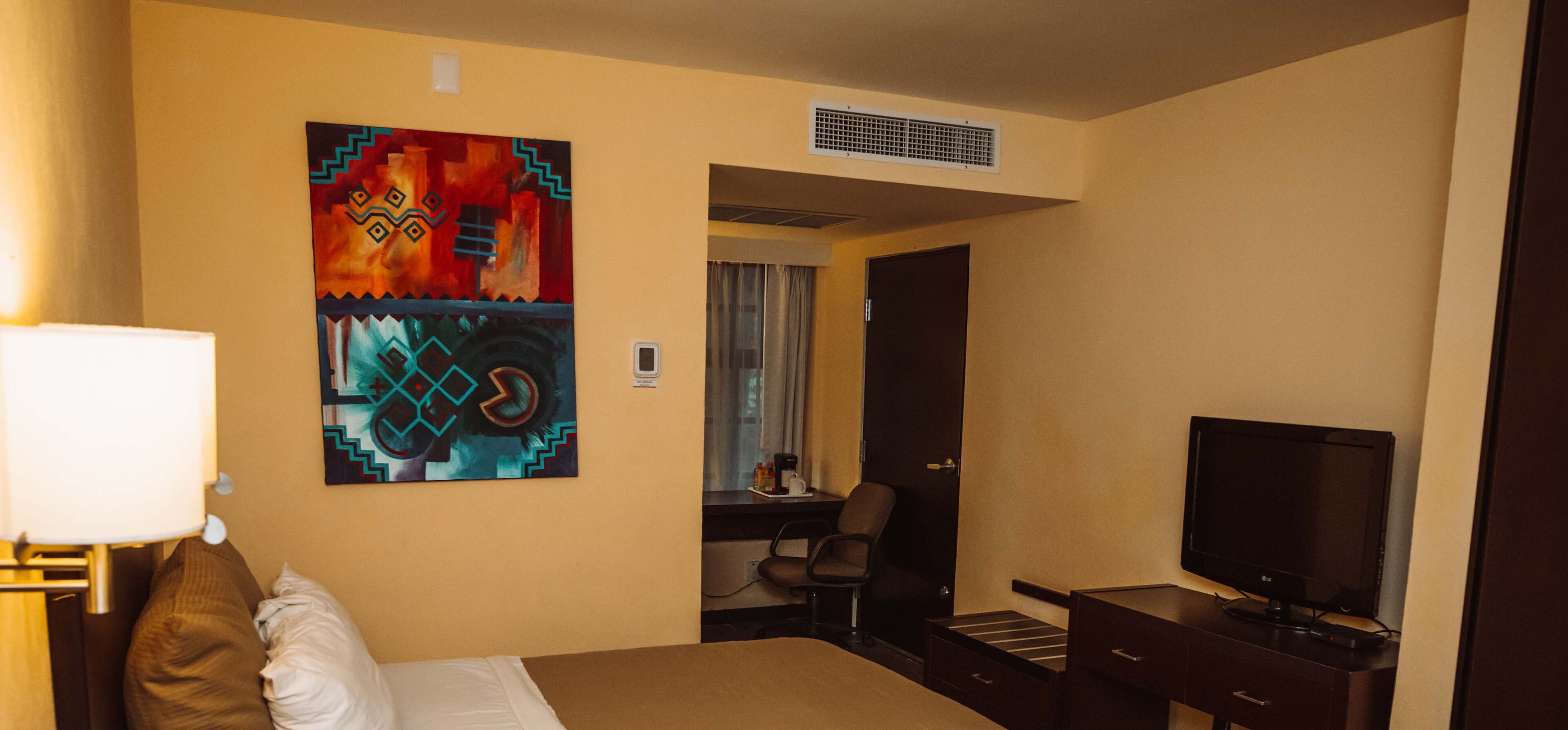 A hotel room with a bed, a desk, a chair, a television, and a painting on the wall.