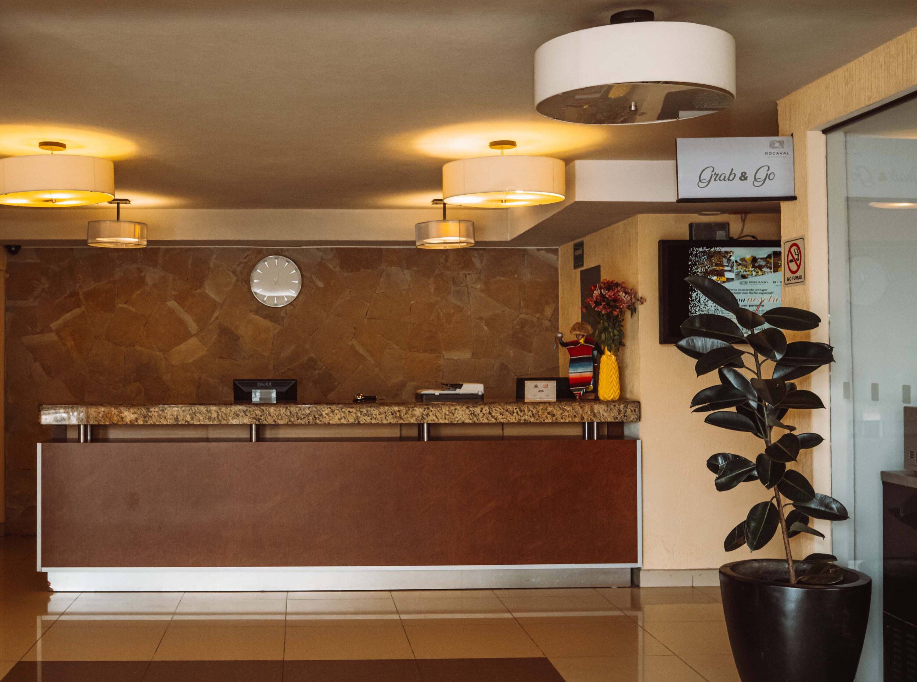 A hotel reception area with a marble wall, a clock, a 'Grab & Go' sign, a reception desk, and a potted plant.