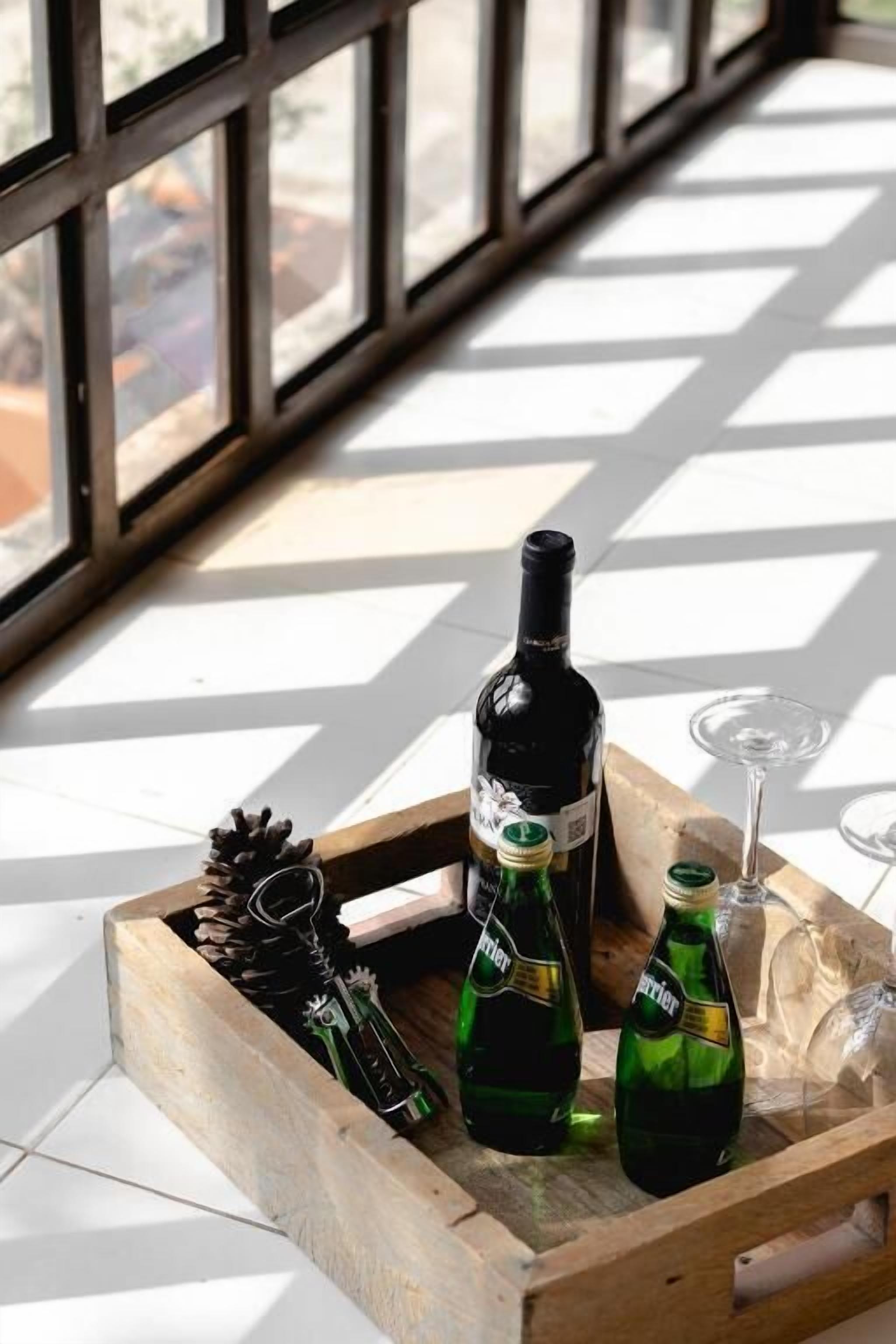 Una bandeja de madera con botellas y una copa de vino sobre un suelo de baldosas, cerca de grandes ventanales.