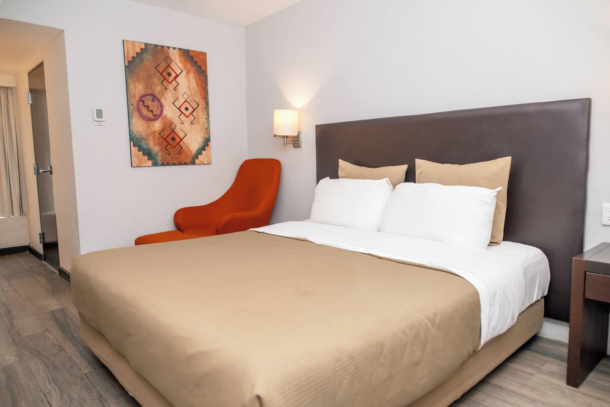 A hotel room with a large bed, a chair, a wall-mounted light, and a painting.