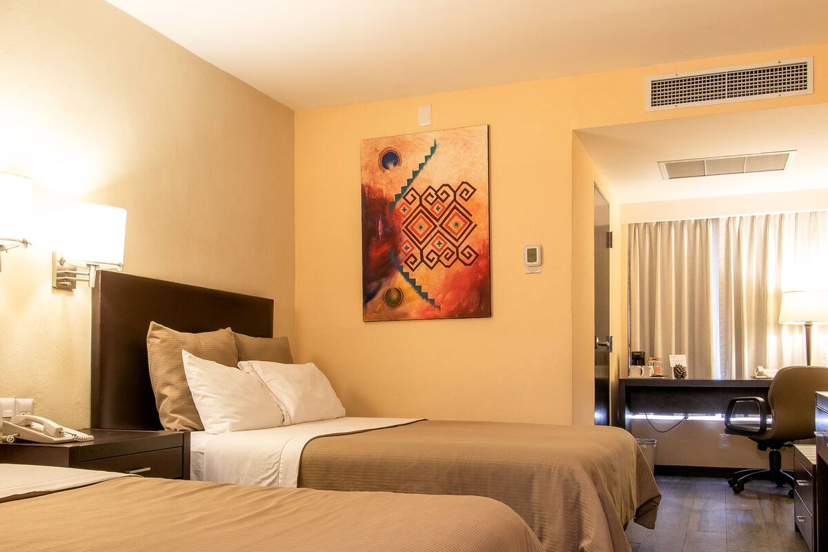 A hotel room with two beds, a desk, a chair, a painting on the wall, and a window with curtains.