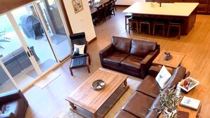Un vaste espace de vie comprenant un grand canapé en cuir marron, une table basse en bois et un coin repas avec une table et des chaises blanches.