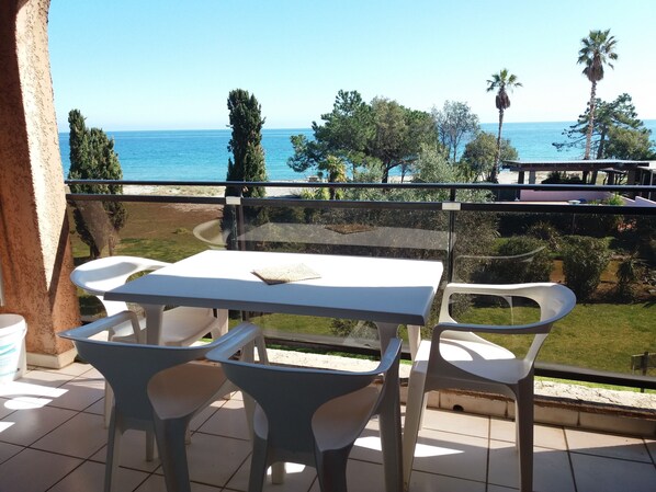 Un balcón con mesa y sillas, con vista al mar, árboles y cielo despejado.