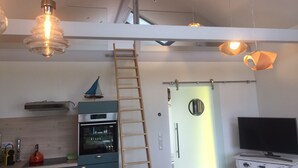 Una cocina moderna con horno empotrado, microondas y hervidor en la encimera. Se ve una escalera de madera que lleva a una zona superior, y un televisor sobre un aparador blanco.