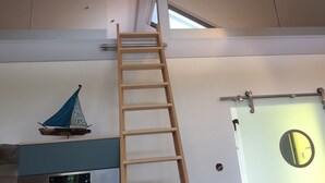 Una habitación con una escalera de madera apoyada en la pared, una pequeña decoración de velero, un espejo circular y una lámpara moderna.