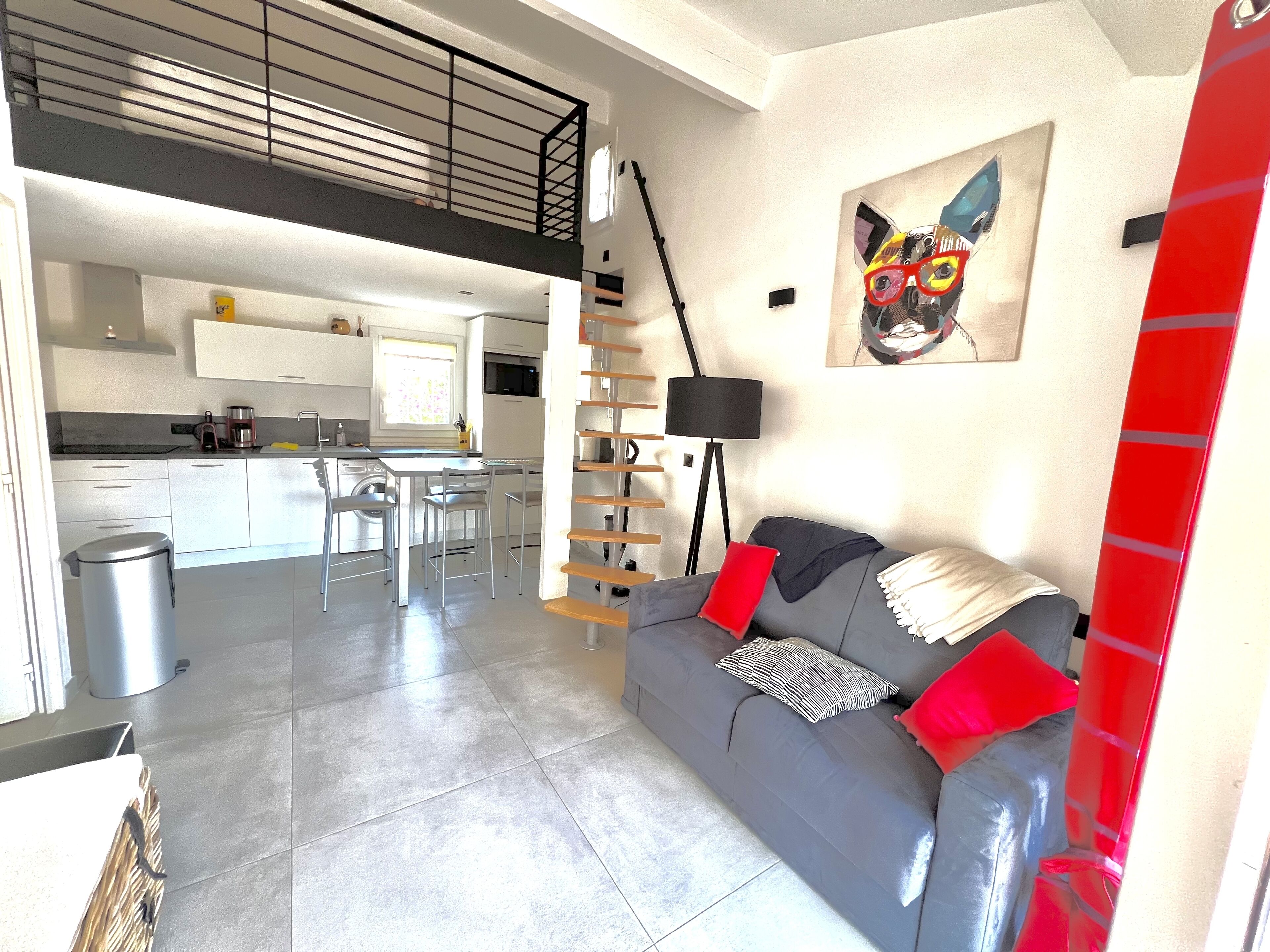 Un espace de vie moderne avec un canapé gris, des coussins rouges, un lampadaire noir et un coin cuisine à l’arrière-plan.
