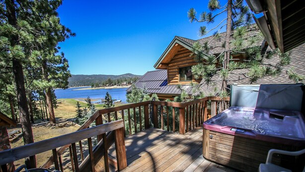 A wooden deck with a hot tub, overlooking a lake and a cabin.