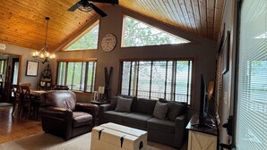 A spacious living room with a high ceiling, wooden beams, a ceiling fan, a large window, a sofa set, a coffee table, and a dining area with a table and chairs. - Lakefront Family Cabin + Sandy Swim Beach + Bonfires + Sunsets+pontoon rental (Minong)
