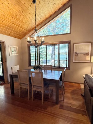 Un coin repas avec une table et des chaises en bois, un lustre, de grandes fenêtres et un tableau encadré au mur.
