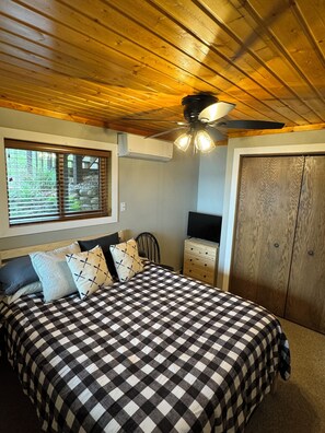 Een slaapkamer met een geruit beddegoed, houten plafond, plafondventilator, een dressoir en een kledingkast.