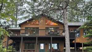 A two-story lodge with a balcony, a stone wall, and a wooden deck. - Lakefront Family Cabin + Sandy Swim Beach + Bonfires + Sunsets+pontoon rental (Minong)