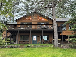 A two-story lodge with a balcony, a stone wall, and a wooden deck. - Lakefront Family Cabin + Sandy Swim Beach + Bonfires + Sunsets+pontoon rental (Minong)
