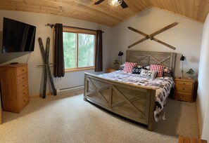 Un dormitorio con techo de madera, una cama con cubrecama escocés, un televisor montado y una ventana con cortinas.
