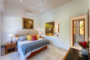 A bedroom with a bed, bedside tables, a ceiling fan, an air conditioning unit, a painting, a mirror, and a vase with flowers. - Villa Cascada Luxury Riverfront Retreat, Villa Cascada Riverfront Pickleball Resort (Puerto Vallarta)
