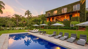 A swimming pool with lounge chairs and umbrellas, surrounded by lush greenery and a house with a green exterior.