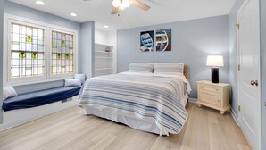 Een slaapkamer met een bed, een nachtkastje, een plafondventilator, een raam met glas-in-lood en een wand met kunstwerken.