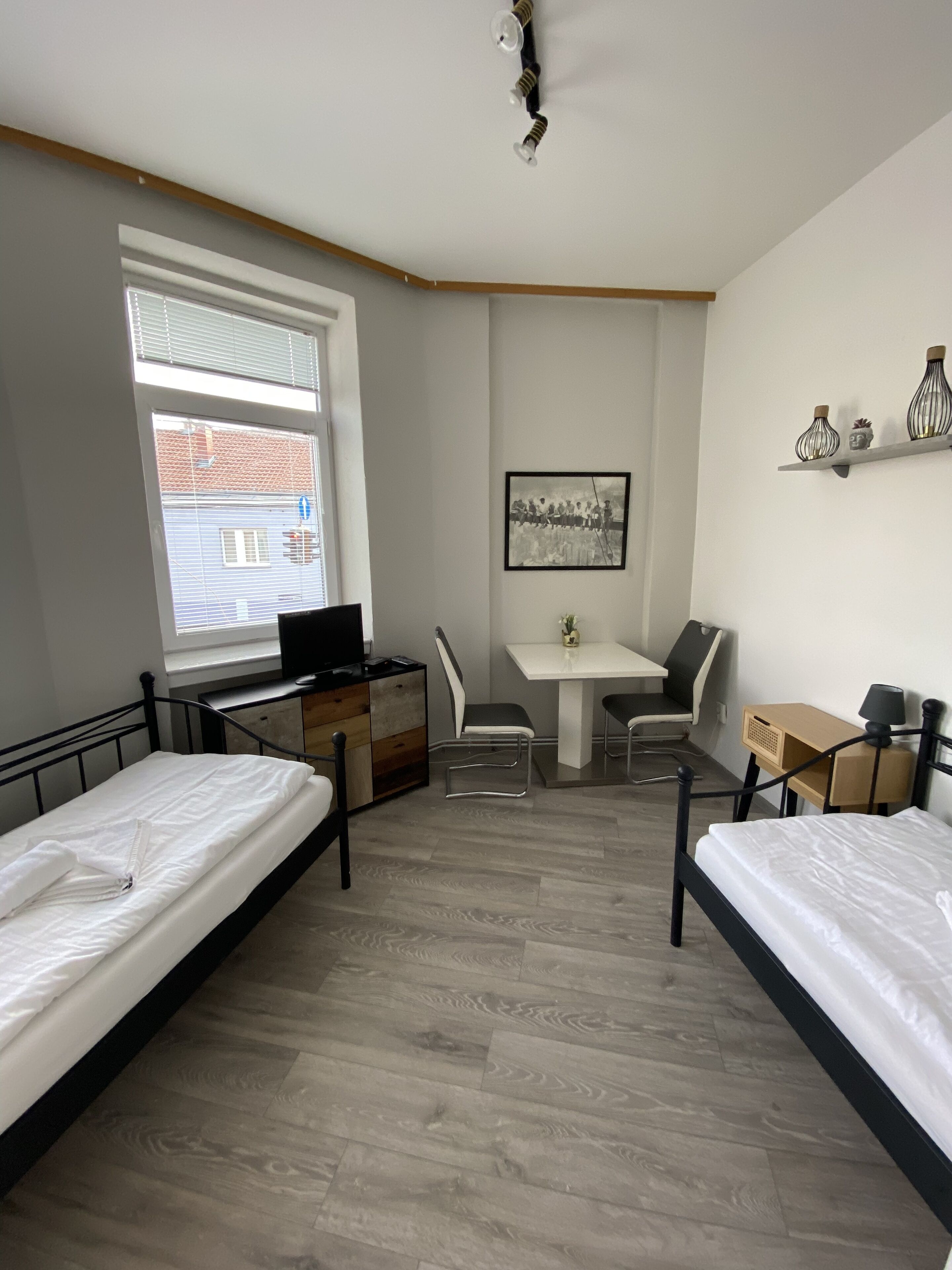 A room with two beds, a desk, a chair, a small table, and a window with blinds.