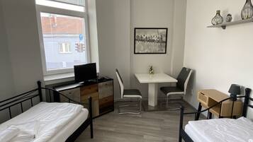 A room with two beds, a desk, a chair, a small table, and a window with blinds.