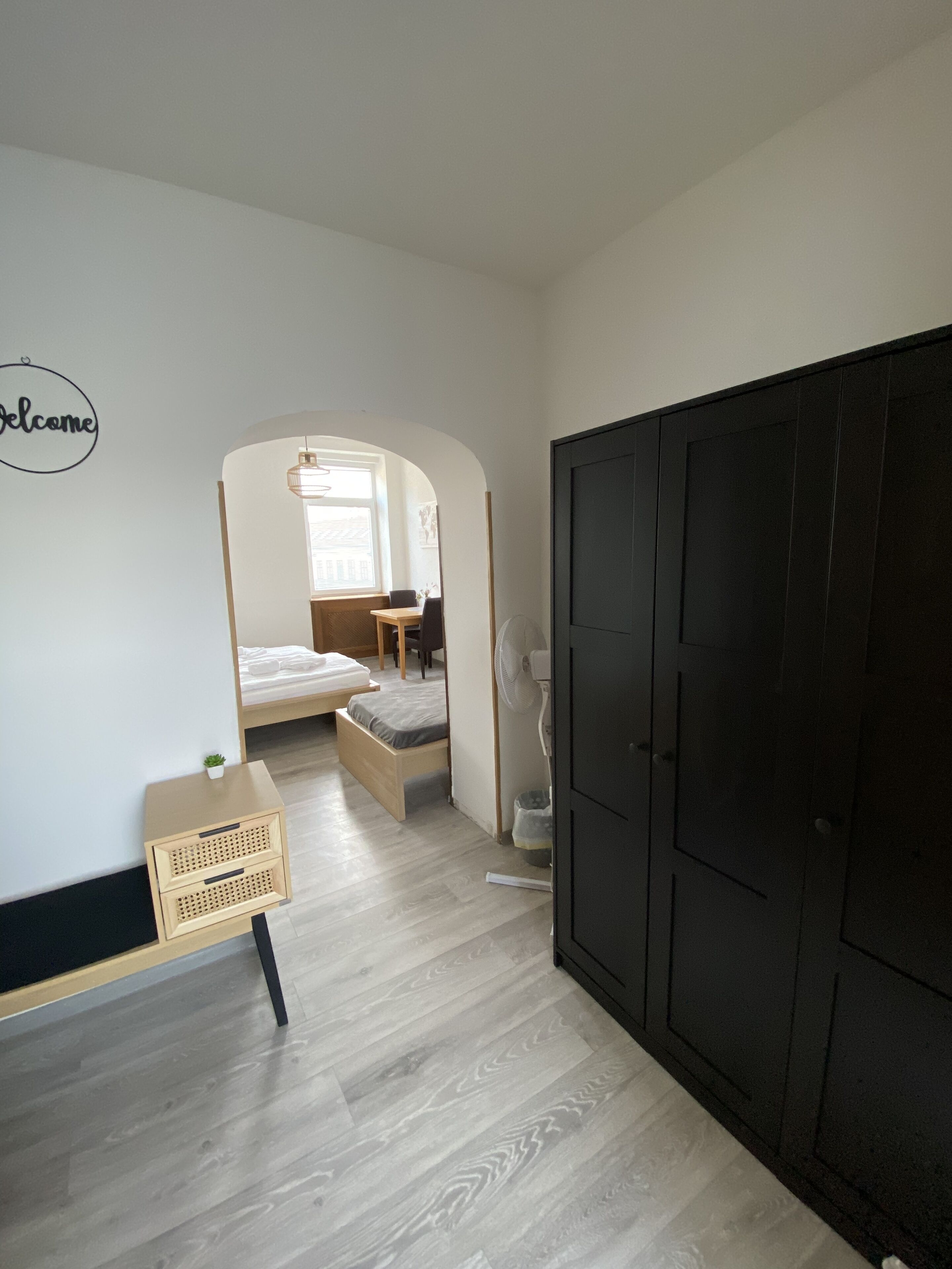 A room with a bed, a small table, a chair, a fan, and a wardrobe.