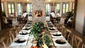 Une longue table à manger, dressée pour un repas, avec des chaises de chaque côté, une cheminée en pierre en arrière-plan et une vue sur l’extérieur grâce à de grandes fenêtres.